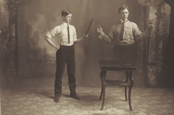 William R. Holmes, member of the Menomonie High School class of 1905, depicted as a professional magician in front of a painted backdrop. Pictured with a magic wand that resembles the leg of a chair, causing an assistant to disappear.