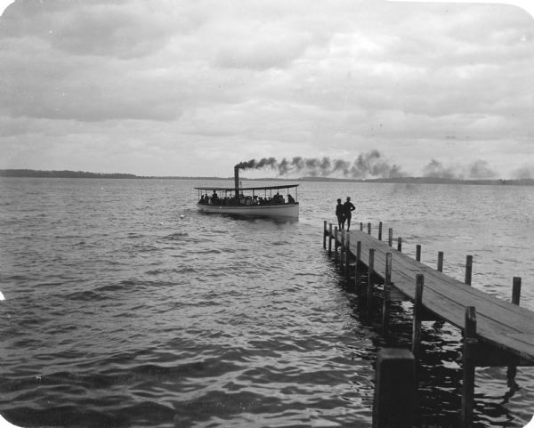 The lake steamer "Alice" approaches the Madison dock on Lake Monona. Two boys wearing caps stand at the end of the pier.