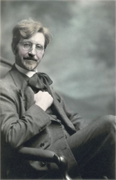 A seated self-portrait of the photographer, Ephraim Burt Trimpey. He wears eyeglasses, and a suit with vest and floppy cravat.