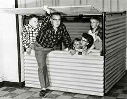 A middle-aged man stepping into small metal crate shelter while holding up a hinged upper door. His wife and four kids are inside the shelter.