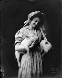 Young woman poses in a dress and bonnet in a studio. She is holding two young pigs.