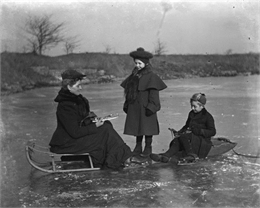 Florentina, Jennie, and Edgar Krueger prepare for ice skating on the quarry pond. Florentina and Edgar sit on sleds on the ice, as they attach blades to their shoes. Jennie stands between them.