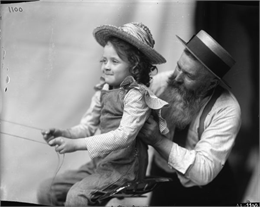 A young girl sits in the seat of a farm implement and holds onto reins as an older bearded man assists. The image was likely used as a model for advertising art.