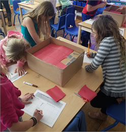 Numerous children looking down as they color and create with red and white paper on a wooden table.
