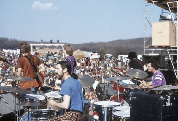 Side view of the Grateful Dead on stage, featuring Bob Weir, Mickey Hart, Phil Lesh, Jerry Garcia, and Bill Kreutzmann, play on stage at the Sound Storm Festival. In the background is a large crowd and tents.