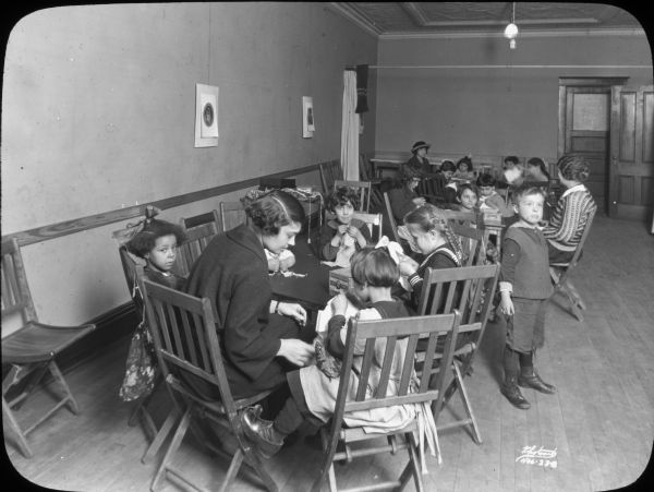 Groups of children seated at tables for a Saturday morning sewing group at Neighborhood House, with a woman helping the children at each table.