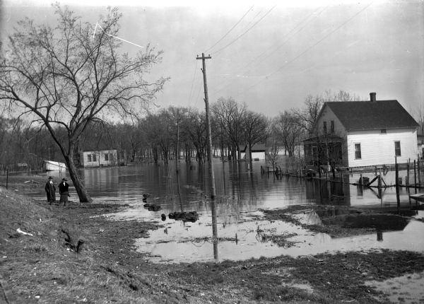 A flood of water has crept up to the edges of two buildings. Two women stand near a tree at the edge of the water.