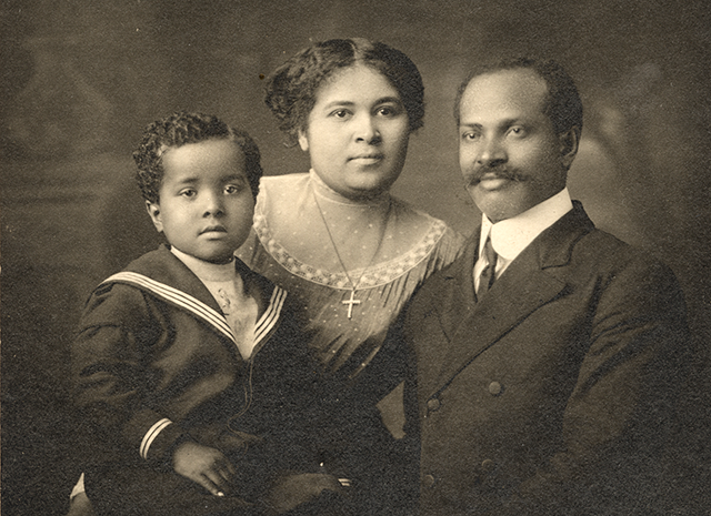 The man is wearing a double-breasted suit, shirt, and tie, the woman wears a sheer-yoked dress and a cross pendant, and the boy is wearing a sailor suit.