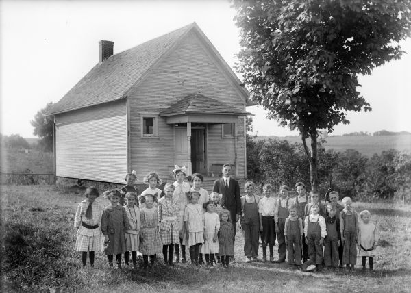 A group of children with their teacher pose on the lawn outside a one-room school building.