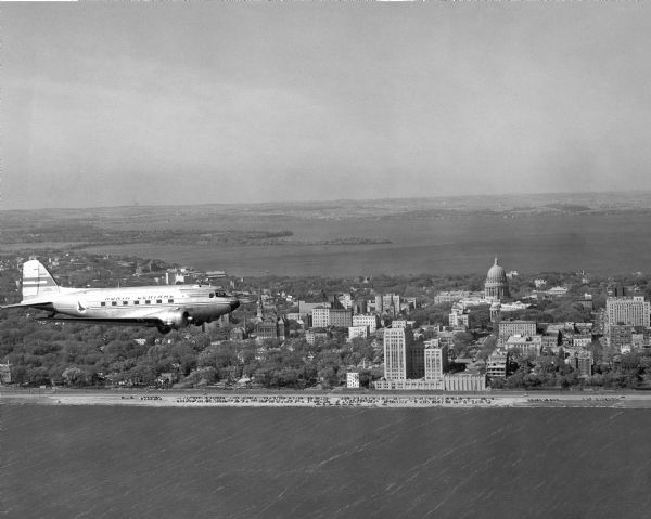 Aerial view of a North Central Airlines DC-3 airplane flying over the Madison isthmus. Clearly visible are the Wisconsin State Capitol, the 1 West Wilson Street State Office Building, and Lakes Monona and Mendota.