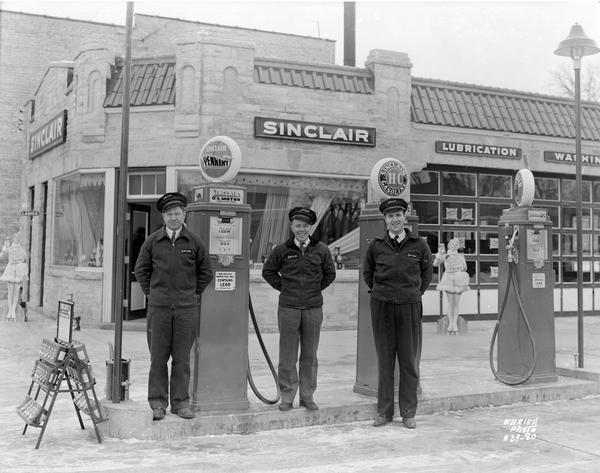 Three uniformed employees of the Sinclair Oil Company Service Station, located at 501 East Broadway, stand in front of the station's gas pumps. Two lifesize cutouts of women are in the background. The station was constructed in 1938.