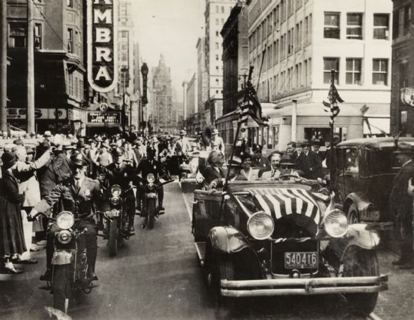 Franklin D. Roosevelt waving from a flag draped car surrounded by policemen on motorcycles.