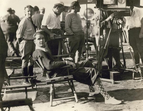 Howard Hughes sitting in a chair among a group of people on the set of 'Sky Devils", a comedy about World War I.