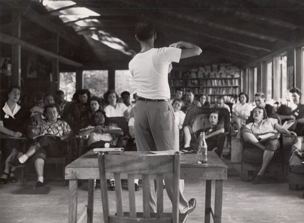 A man, with his back to the camera, is gesturing at and lecturing a student class, who are seated in a variety of chairs.