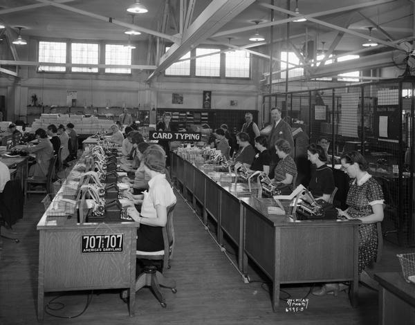 A view of rows of desks with women employees. Behind the work area two men, the supervisor and the Secretary of State Fred Zimmerman, stand.