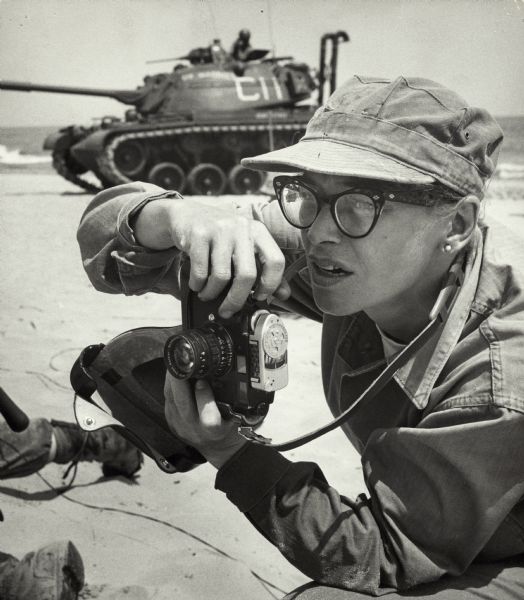 Dickey Chapelle, holding her camera while framing in a shot. There is a tank in the background.