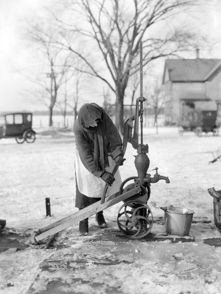 A bundled-up woman hand pumps water from a well into a pail. The ground is covered with ice and snow. Two cars and a farmhouse appear in the background.