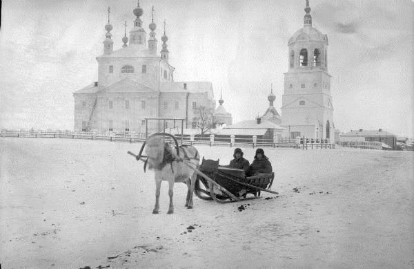 Two officers wearing heavy winter clothes are seated in a horse-drawn wooden sleigh on a snowy path. Behind them is a building that appears to be a Russian church.
