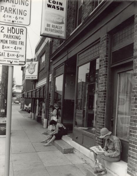 View down a sidewalk towards three people waiting outside of a coin laundromat on Williamson Street. Two women, one on steps of a doorway and another near a closed door, are sitting and reading newspapers. Next door to the laundromat is Star Liquor.