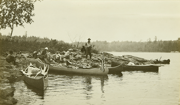 The Gang stopping at Sand Point for a lunch break. The canoes are at the edge of the shoreline, while everyone is sitting on the ground eating.