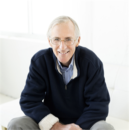 Headshot of author Doug Moe. He is pictured sitting down, wearing glasses, a blue quarter-zip jacket, and grey pants.