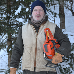 Author James Card wearing a blue beanie hat, camouflage scarf, grey jacket and tan vest. Card is holding an orange chainsaw by his side.