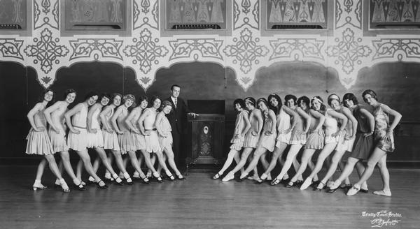 Leo Kehl in a line with 21 female dance students. The young women are posed in a kickline position, and most are wearing skirts, socks and tap or ballroom shoes.