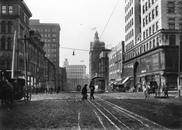 A muddy street in Milwaukee with a streetcar, horse-drawn carts and pedestrians.