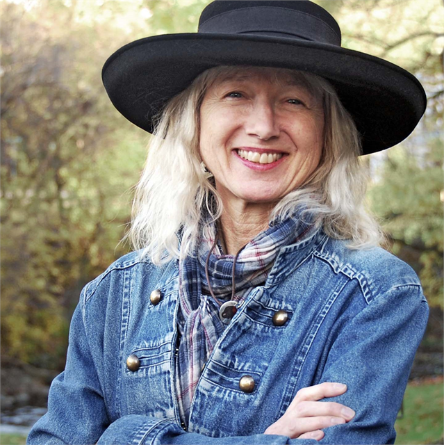 Renée Graef smiles at camera with arms crossed in a black hat, jean jacket, and scarf