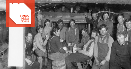 A large group of men, probably logging workers, are shown playing cards in a sleeping shanty.