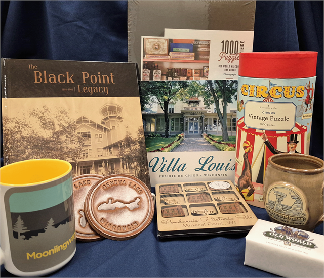 A display of Wisconsin-themed gifts including 'The Black Point Legacy' and 'Villa Louis' books, a Mooningwear mug, Geneva Lake leather coasters, a Wisconsin Dells pottery mug, Old World Wisconsin soap, a Pendarvis Historic Site magnet set, a 1000-piece puzzle of Old World Wisconsin, and a Circus-themed vintage puzzle, all arranged on a dark blue cloth.