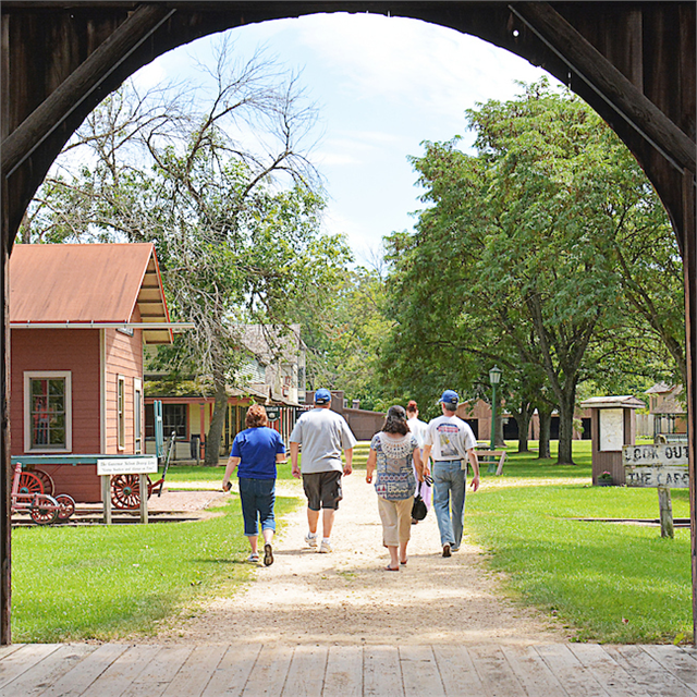 Visitors walking through a historic village area, viewed from inside a wooden covered bridge, with trees and vintage buildings lining the path ahead.