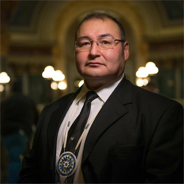 Photograph of Bill Quackenbush, a Native American man wearing wire-rim eyeglasses, a black suite, white shirt, black tie, and Native beaded medallion