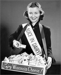 Portrait of Margaret McGuire wearing a beauty contestant sash that says 'Alice in Dairyland' holding a box of cheese.
