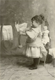 Margery Bish as a little girl wears a striped dress while hanging clothes on a clothesline. There is a doll behind her on the right.