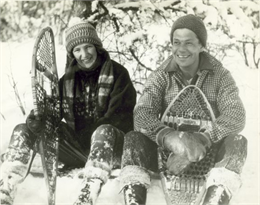 A couple sitting outdoors in the snow holding snowshoes.