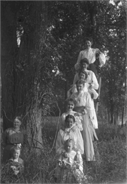 Seven girls and women are arranged on a ladder in the woods. Next to them are two more girls posing in front of a tree.