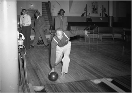 Down-lane view of Isaiah Pyant, member of C.I.O., bowling. A few men are visible in the background walking past, looking on and sitting on a bench.