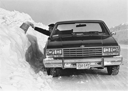 A mail carrier reaches over and almost through a snow bank to put mail into a rural mailbox.