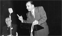 Joseph McCarthy speaks from a podium in front of a large microphone. He is leaning forward and pointing one finger at the audience.
