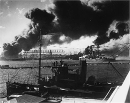 A tug boat in the foreground and a battleship sit in Pearl Harbor with smoke rising.