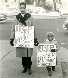 A woman and small child are standing on a sidewalk with 'Stop School Segregation' signs.