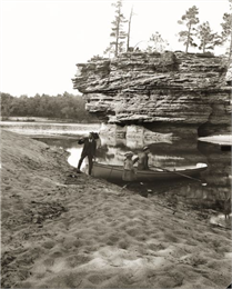 H.H. Bennett and Ruth Bennett on shore near Sugar Bowl. H.H. Bennett holds a camera. Unidentified man in rowboat.