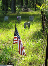Grassy cemetery with four gravestones and an American flag in the foreground.