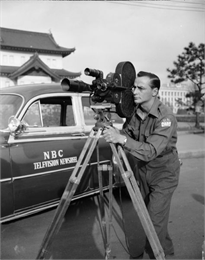 A U.N. war correspondent is shown filming with a news camera next to an NBC Television Newsreel vehicle.