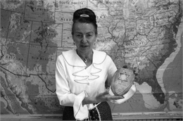 Magda Herzberger stands in front of a map of the United States holding a wooden plaque carved with a Star of David and the inscription, "In Gratitude to Magda Herzberger."