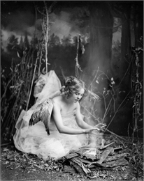 A model posed as an angel sits on the ground in front of a sylvan backdrop, warming her hands over a fire. She wears wings and a dress of diaphanous material.