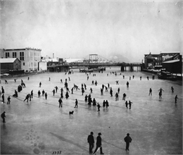 Elevated view of a large group of people skating on the frozen Manitowoc River between the 8th and 10th Street bridges.