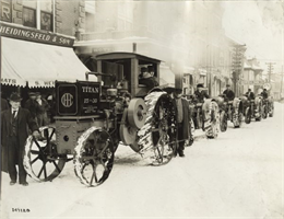 Several tractors, including a Titan 15-30, lined up along a curb near storefronts on a snow-covered Washington Street in downtown Greenfield.