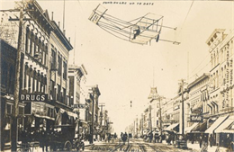 Street scene in Fond du Lac featuring many pedestrians, horse-drawn carriages, shops, and other buildings. An image of a plane has been added to the sky.